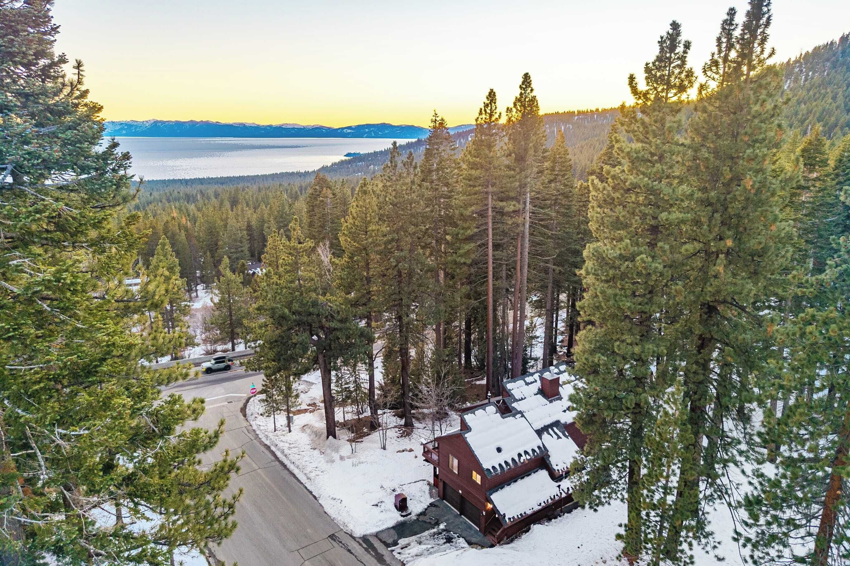 A cabin surrounded by tall pine trees, a road, and a lake with mountains in the background.