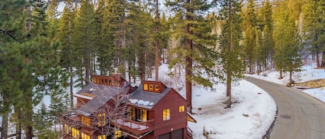 A two-story cabin with a garage, surrounded by tall pine trees and a snow-covered driveway.