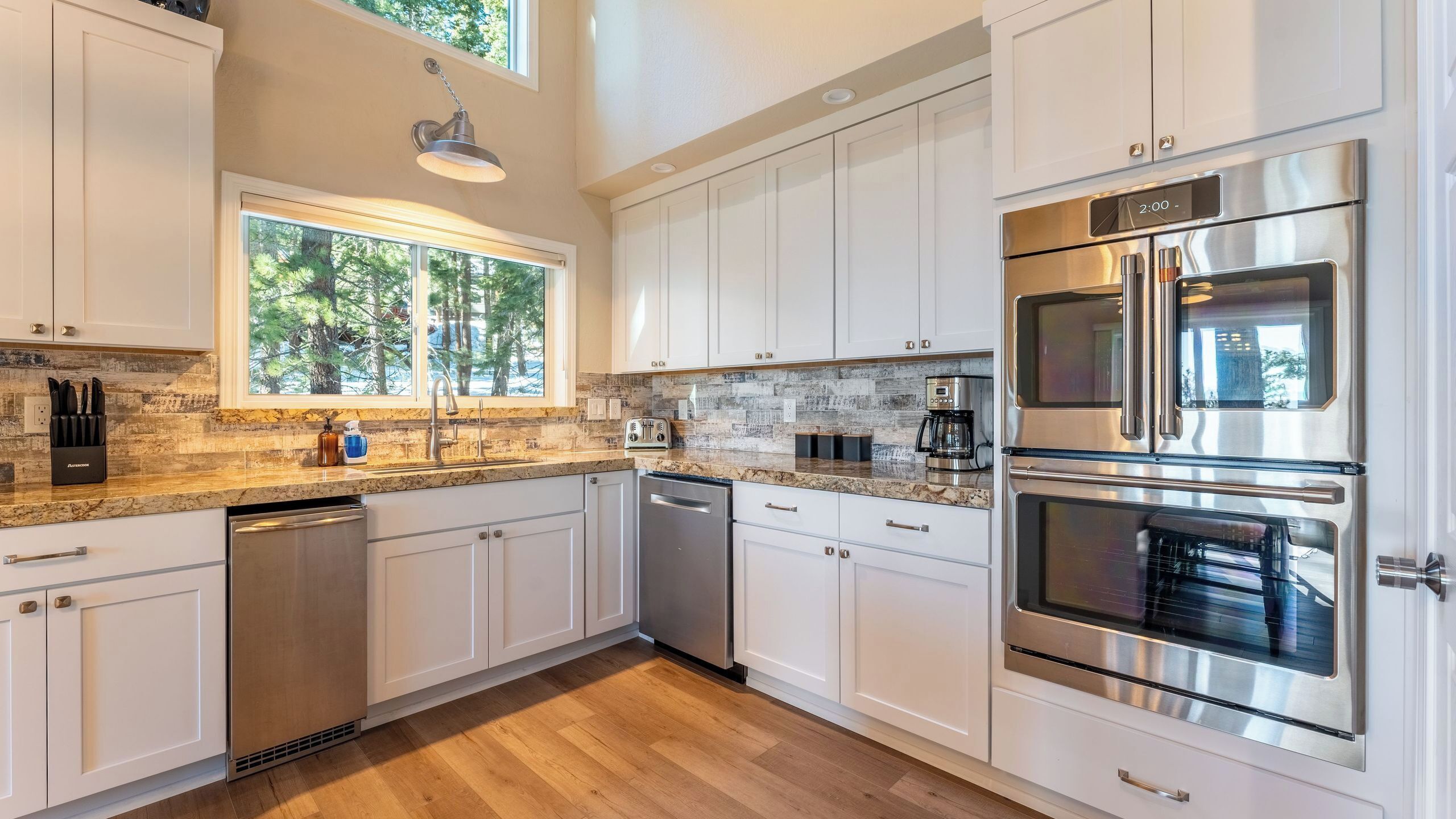A modern kitchen with white cabinets, stainless steel appliances, a window with a view of trees, and a built-in oven.