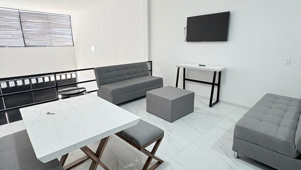 A modern living room with a white sofa, grey ottoman, and a white coffee table with wooden legs. - Periférico Bodega con Departamento C (Tonalá)