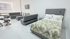 A modern hotel room with a large bed, grey sofas, a small table, and a window with blinds. - Periférico Bodega con Departamento C (Tonalá)
