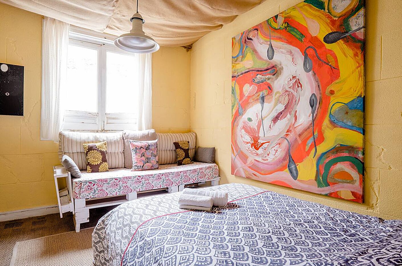 Un dormitorio con cama, ventana con cortinas, banco con almohadones y un cuadro abstracto grande en la pared.