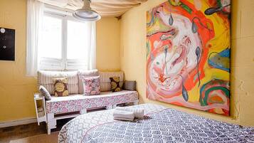 Un dormitorio con cama, ventana con cortinas, banco con almohadones y un cuadro abstracto grande en la pared.