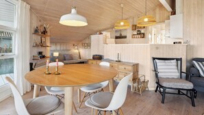 A wooden interior with a round dining table, white chairs, a black chair with a striped cushion, and pendant lights.