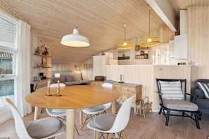 A wooden interior with a round dining table, white chairs, a black chair with a striped cushion, and pendant lights.