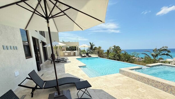 A poolside area with lounge chairs and umbrellas, a clear view of the ocean, and a modern building with large windows. - White Sky Beach Villas (New Hope)
