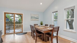 Uma sala de jantar com mesa de madeira posta para seis lugares, rodeada por cadeiras, com vista para um deck e mobiliário exterior.