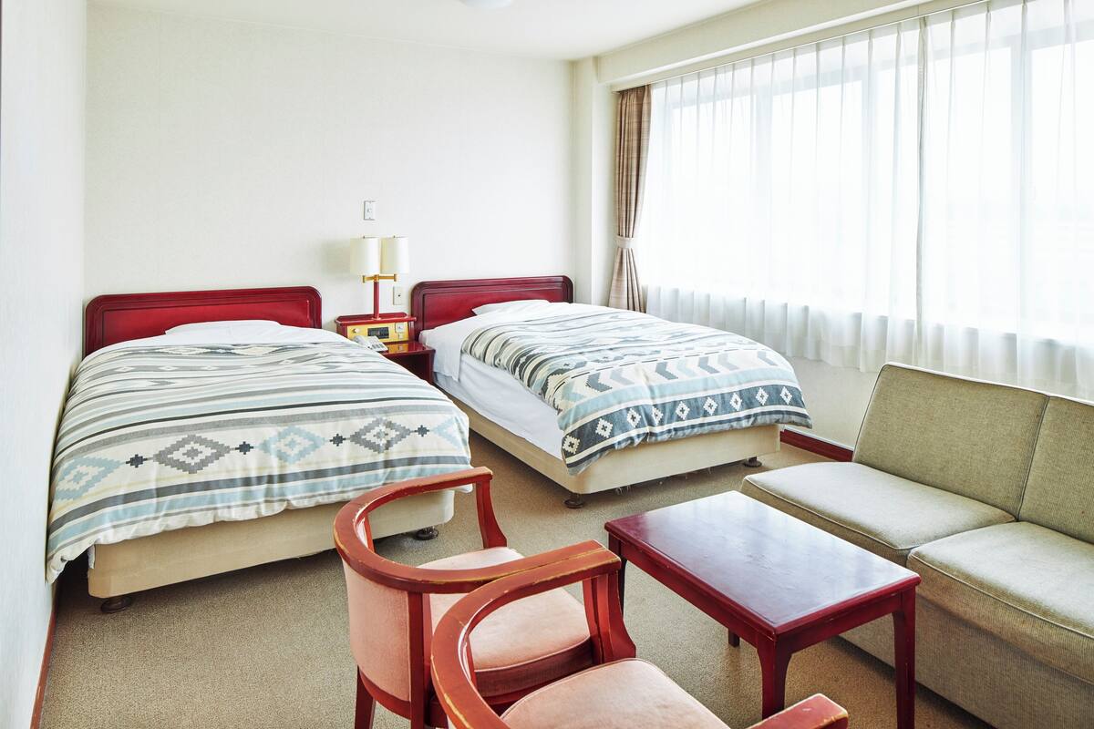 A hotel room with two beds, a sofa, a chair, a small table, and a nightstand with a lamp.