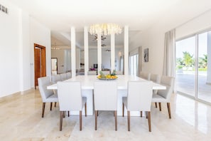 A modern dining area with a white table, grey chairs, a chandelier, and a large window with a view of palm trees. - Modern ocean view villa with chef at Cap Cana, an exclusive Golf & Beach Resort. (Punta Cana)