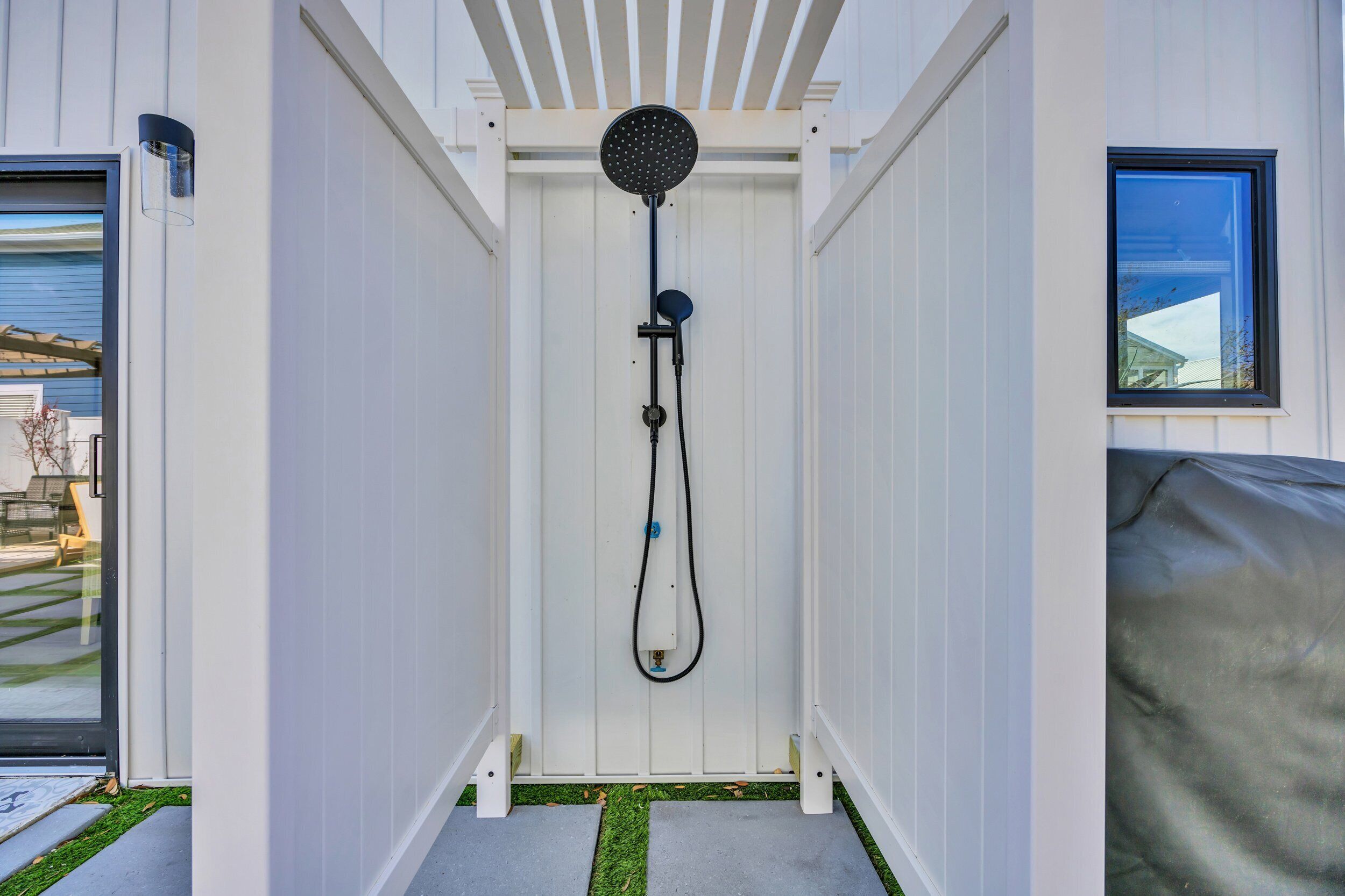 Une douche extérieure moderne, dotée d’un pommeau de douche à effet pluie, entourée de murs blancs et agrémentée d’une petite fenêtre sur le côté droit.