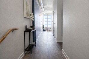Ein Korridor mit Geländer aus Holz, einem Konsolentisch mit Vase und Pflanze, und Blick in einen Raum mit Stuhl und Fenster.