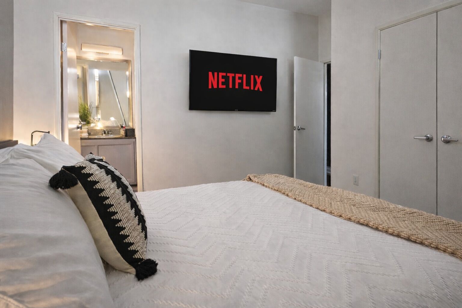 Ein Schlafzimmer mit einem Bett, einem an der Wand montierten Fernseher, der 'NETFLIX' zeigt, einem Kleiderschrank und einer Tür zu einem Badezimmer.