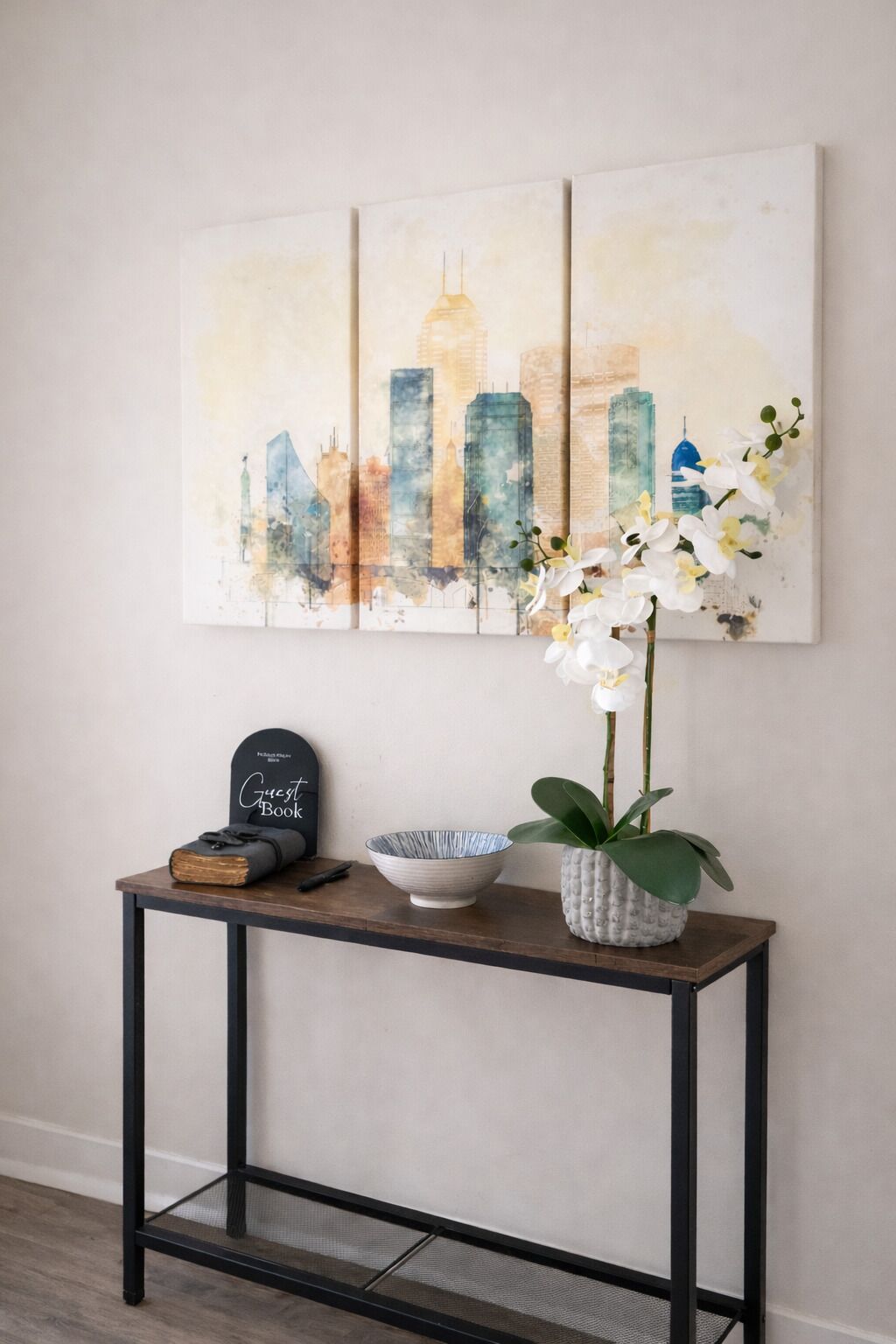 Una obra de arte con una vista urbana, un libro de visitas, un cuenco y una orquídea sobre una consola de madera.