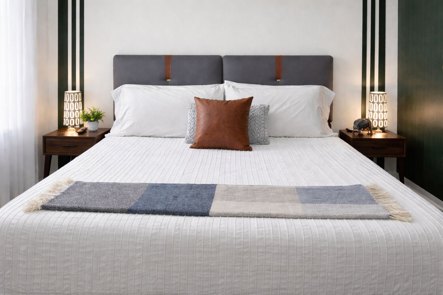 A bed with a white quilt, two pillows, a brown throw, and a blue and white area rug. There are two bedside tables with lamps and a small plant on one.