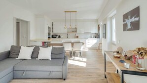 Un saló modern amb un sofà gris, una zona de menjador amb cadires blanques, una cuina amb armaris blancs i il·luminació penjant.