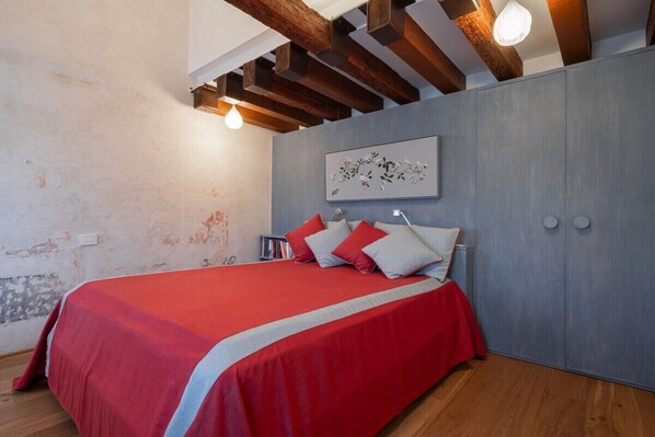 A bedroom with a bed, wooden beams, a light fixture, a wall-mounted artwork, and a wardrobe.