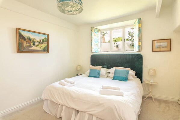 A neatly made bed with white bedding and blue pillows, a painting on the wall, and a window with curtains.