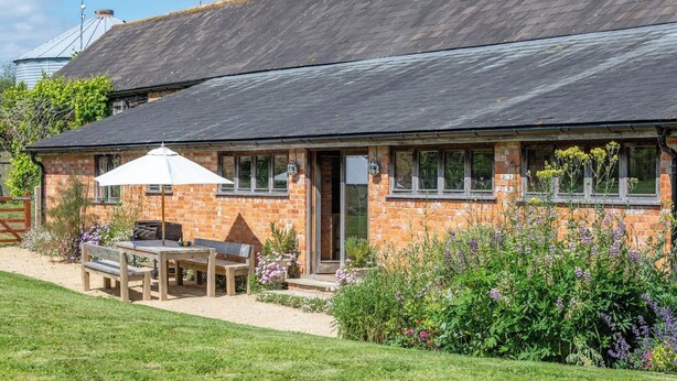 A brick house with a sloped roof, a patio with a table and chairs, and a garden with various plants. - Battys Barn at Moorbath Farm (Bridport)