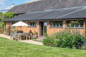 A brick house with a sloped roof, a patio with a table and chairs, and a garden with various plants. - Battys Barn at Moorbath Farm (Bridport)