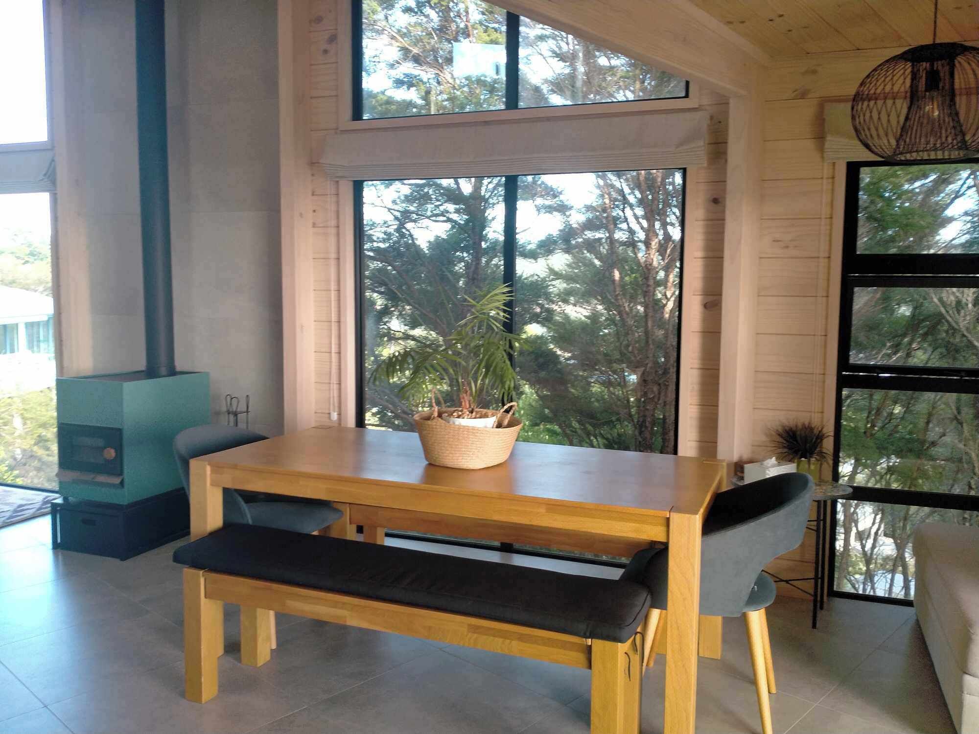 Een houten eettafel met een bankje, een potplant en een groot raam met uitzicht op bomen.
