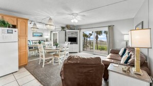 A modern living room with a brown leather sofa, a dining area with white chairs, a kitchen with white appliances, and a balcony with a view of palm trees. - Oceanfront, 2nd Floor, Sea View, 2 Pools (Panama City Beach)
