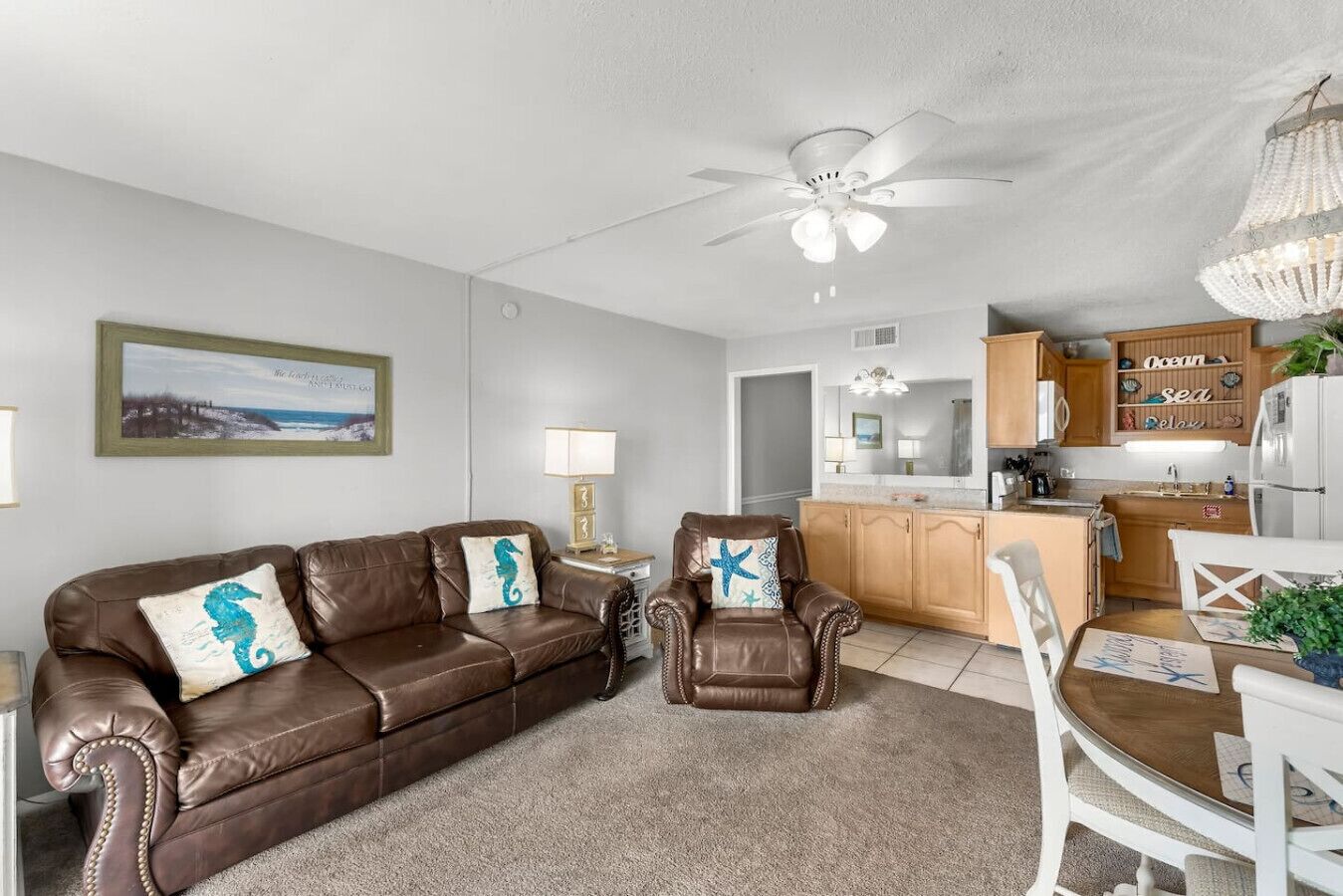 A living room with a brown leather sofa, a brown armchair, and a dining table with white chairs. There is a ceiling fan, a chandelier, and a kitchen area with wooden cabinets and a refrigerator.