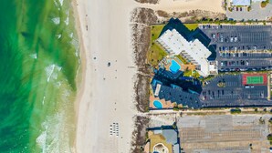 An aerial view of a coastal hotel with a pool, parking lot, and beach access.