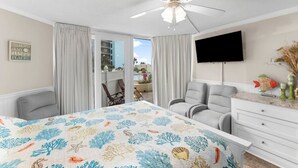 A hotel room with a bed, two armchairs, a TV, and a view of the outdoors.