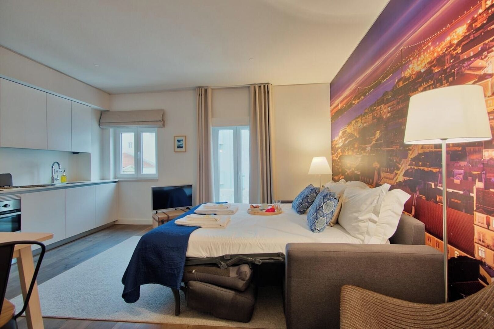 Hotellihuone, jossa on sänky, työpöytä, tuoli, sohva, televisio ja seinällä kaupunkimaisemaa esittävä seinämaalaus.