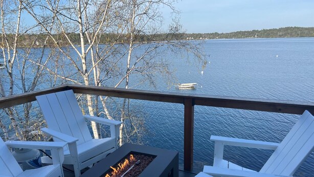 En terrass med vita Adirondack-stolar och en inbyggd eldstad med utsikt över en sjö.