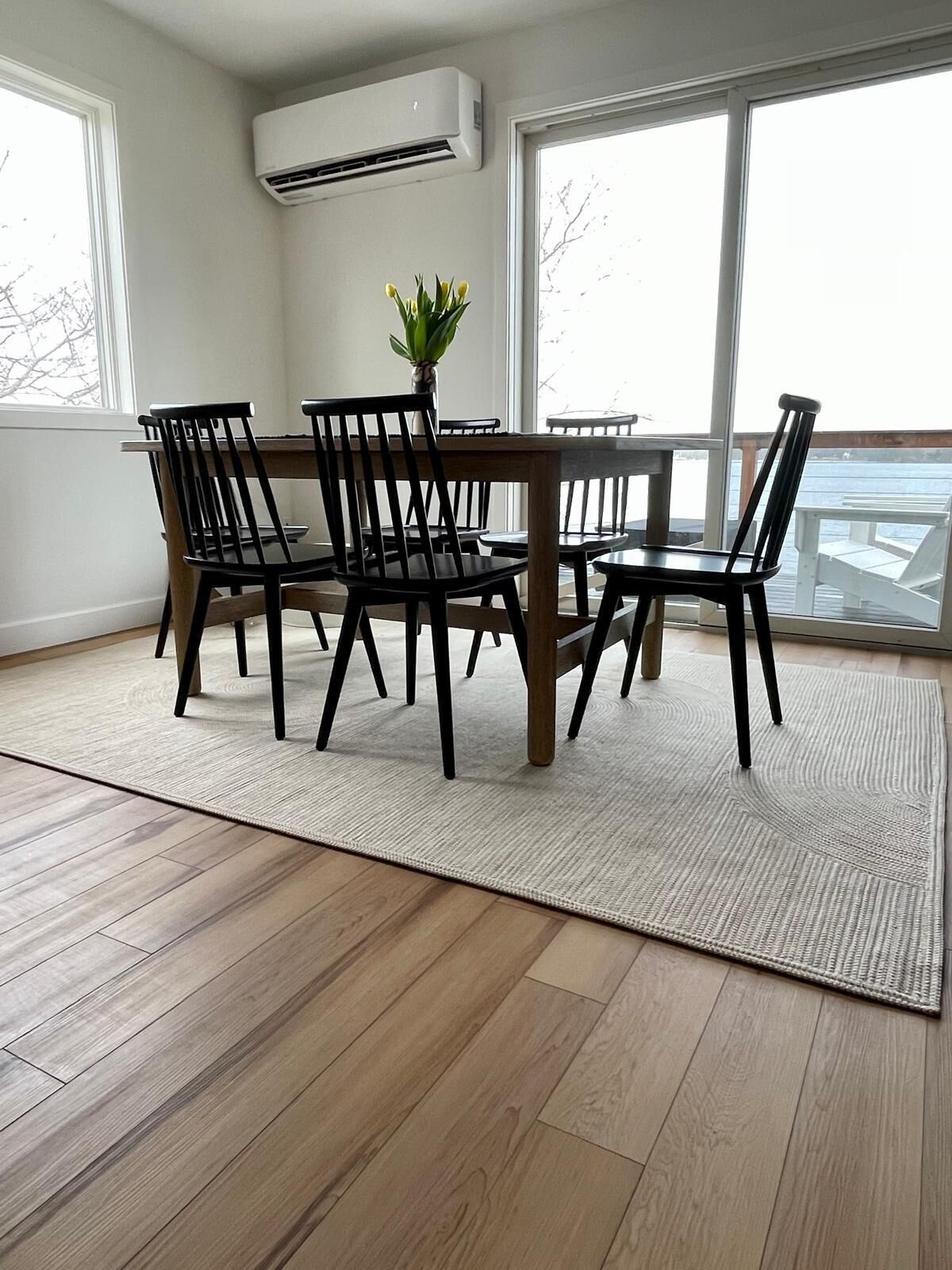 Et spiseområde med et træbord og stole, en potteplante og store vinduer med udsigt til det fri.