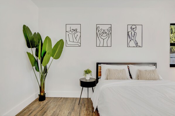 Une chambre à coucher avec un lit, une petite table ronde, une plante en pot et trois œuvres d’art réalisées au trait, accrochées au mur.