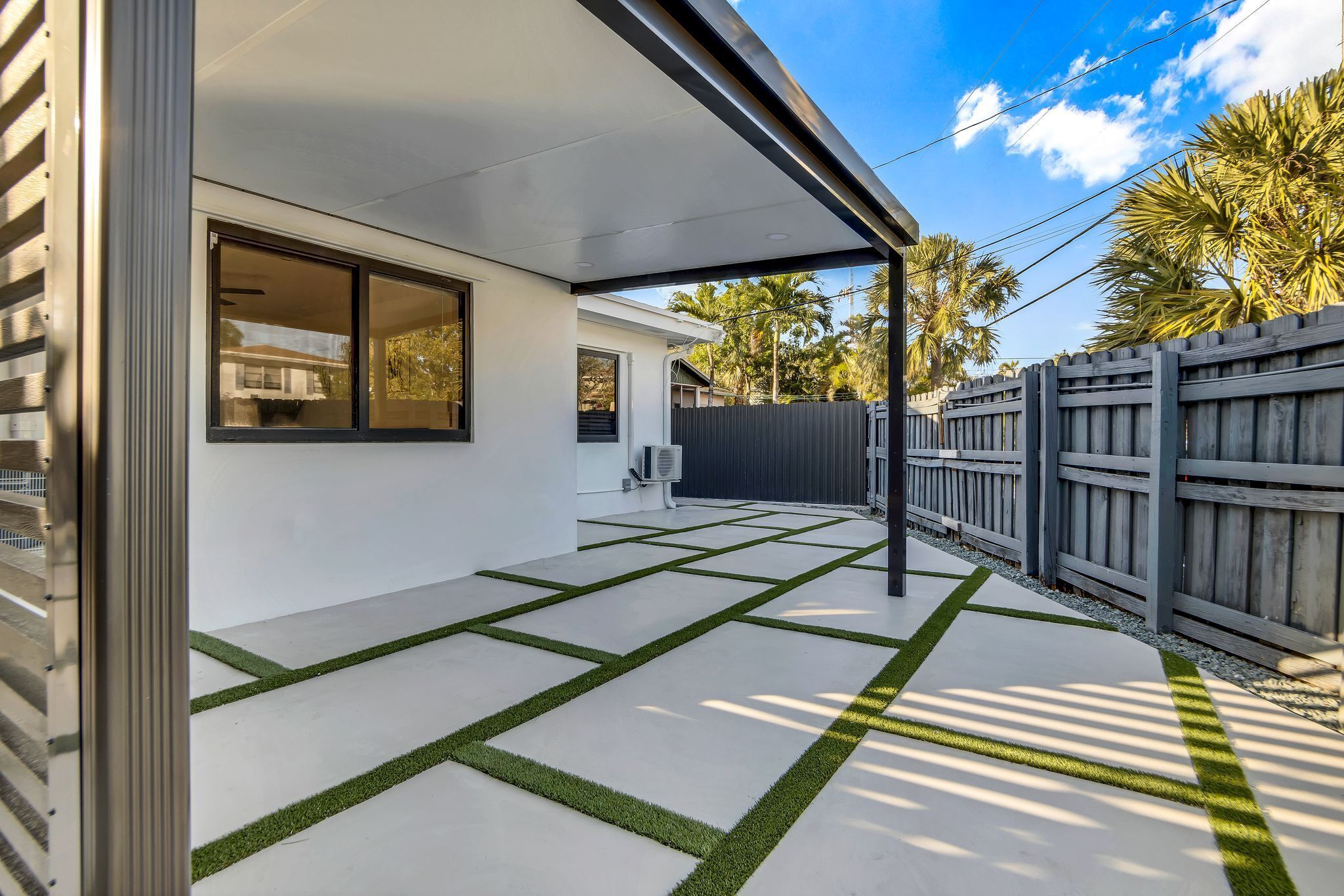 Een moderne patio met betonnen vloer en groene kunstgrasranden, een houten schutting en een heldere lucht.