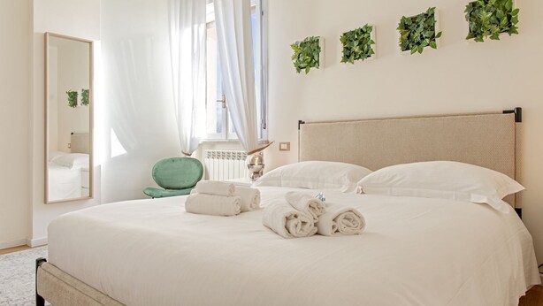 A neatly made bed with white linens and rolled towels, a green chair, a mirror, and wall-mounted greenery. - Our newly renovated property is designed to offer you maximum comfort. The bright and spacious bedroom is equipped with a comfortable double bed, ideal for a regenerating sleep. The living room is perfect for relaxing, with a sofa that turns into a bed t (Pisa)
