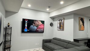 A modern living room with a sectional sofa, a flat-screen TV mounted on the wall, a small shelf with decorative items, and a patterned rug on the floor. - Charming 1-Bedroom Basement Walkout Suite with a Gym and Office. (Calgary)