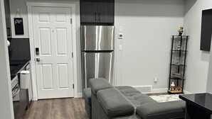 A compact kitchen with a stainless steel refrigerator, a grey cushioned bench, and a small bar stool. - Charming 1-Bedroom Basement Walkout Suite with a Gym and Office. (Calgary)