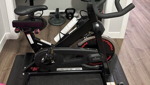 A stationary bike in a room with two black chairs, a weight plate, and a scale. - Charming 1-Bedroom Basement Walkout Suite with a Gym and Office. (Calgary)