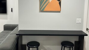 A modern kitchen with a black countertop, two black bar stools, and a wall-mounted artwork of a woman's profile. - Charming 1-Bedroom Basement Walkout Suite with a Gym and Office. (Calgary)