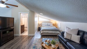 A modern bedroom with a sloped ceiling, a large bed, a TV, and a cozy seating area with a leather couch and a small table. - 4BR Mountain Retreat , Fireplace, Deck with Views, RMNP (Fraser)
