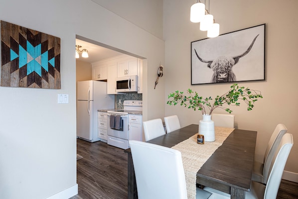 A modern kitchen with white cabinets, a microwave, and a stove. There is a dining table with white chairs and a vase with green plants.