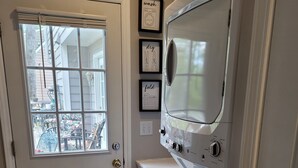A laundry room with a front-loading washer and dryer, a window with blinds, and three framed pictures on the wall. - Charming 1-bedroom apartment in cool Garner, minutes from Raleigh (Garner)