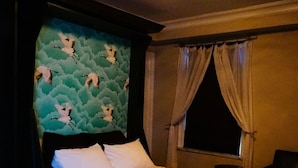Hotellihuone, jossa on sänky, kaksi yöpöytää, lintukuviollinen pääty, ikkuna verhoilla ja kattovalaisin.