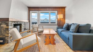 A living room with a stone fireplace, a blue sofa, a wooden coffee table, and a view of mountains through large windows. - Eden Roc 6 - 4 pers - près des pistes (Haute-Nendaz)