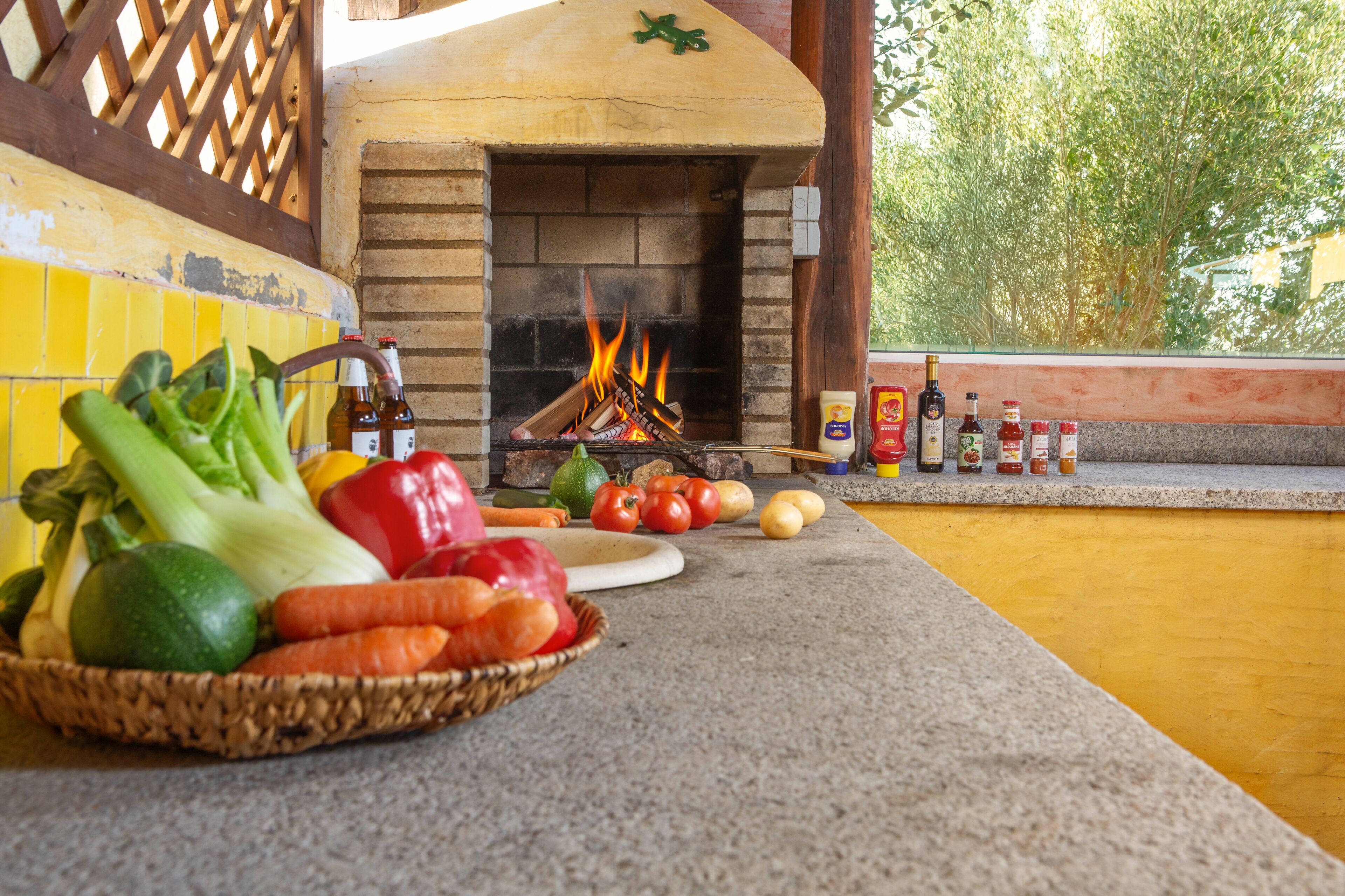 A brick oven with fire, surrounded by vegetables and condiments.