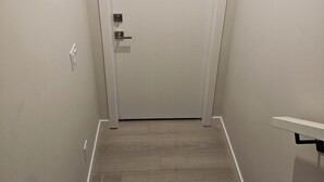Un pasillo estrecho con una puerta blanca al final, suelo de madera y un estante blanco en la pared derecha.