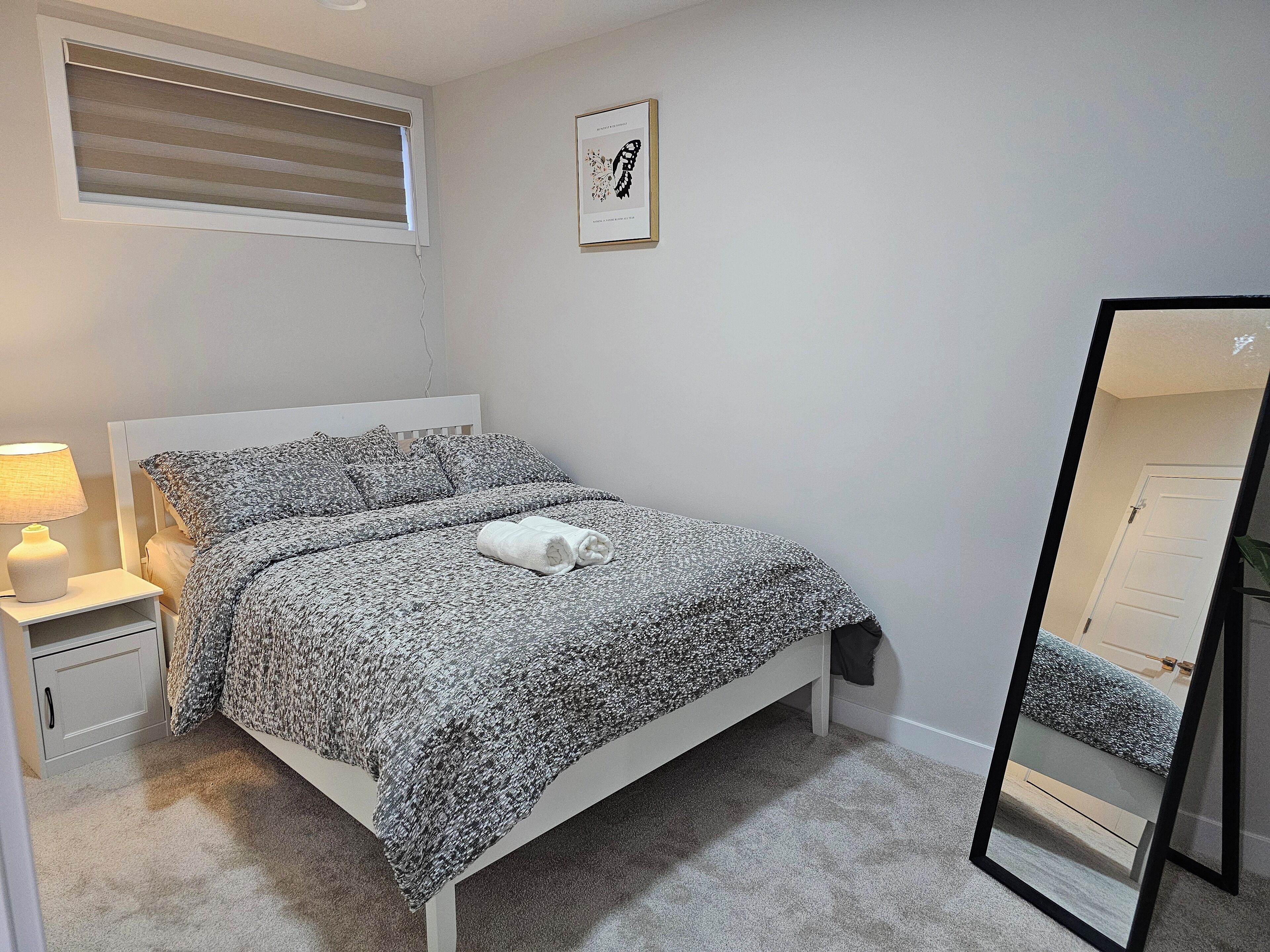 Een slaapkamer met een bed, een nachtkastje met een lamp, een ingelijst beeld aan de muur en een spiegel.