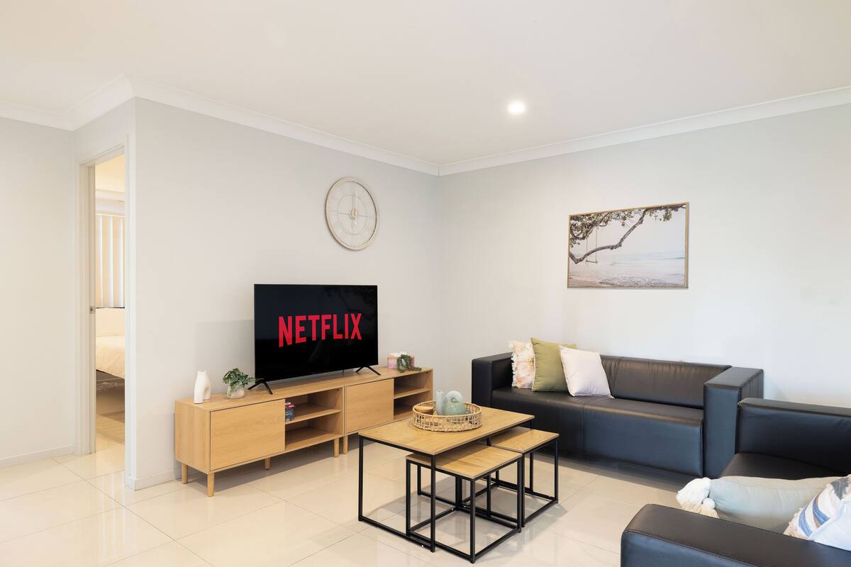 A modern living room with a TV displaying "NETFLIX," a wooden coffee table, a black leather sofa, and a painting on the wall.