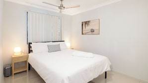 Un lit bien fait, avec du linge de lit blanc, une lampe de chevet et un ventilateur de plafond.