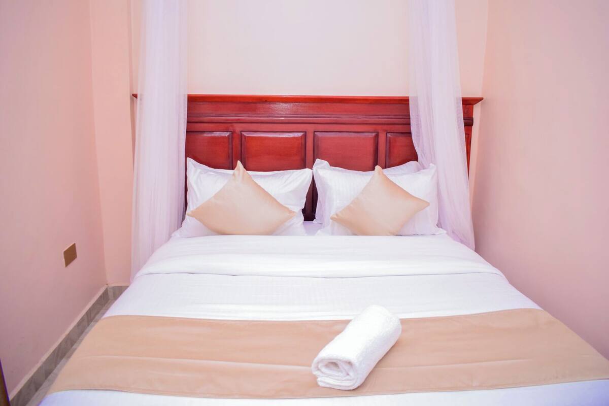 Una cama bien hecha con sábanas blancas, una manta beige y una toalla enrollada.