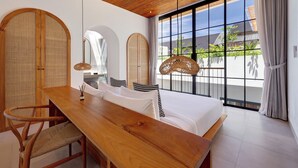 A modern hotel room with a wooden ceiling, large windows with a view of greenery, a wooden dining table, and a bed with white linens. - Villa Kala (Canggu)